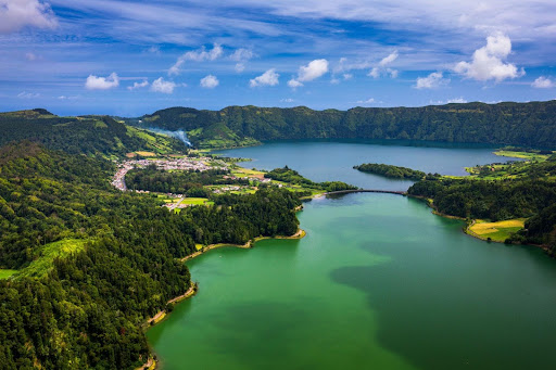 Lagoa das Sete Cidades View of Lagoa das Sete Cidades in the Azores, with two green-blue lakes surrounded by rolling hills, forested slopes, and a small village along the shoreline.